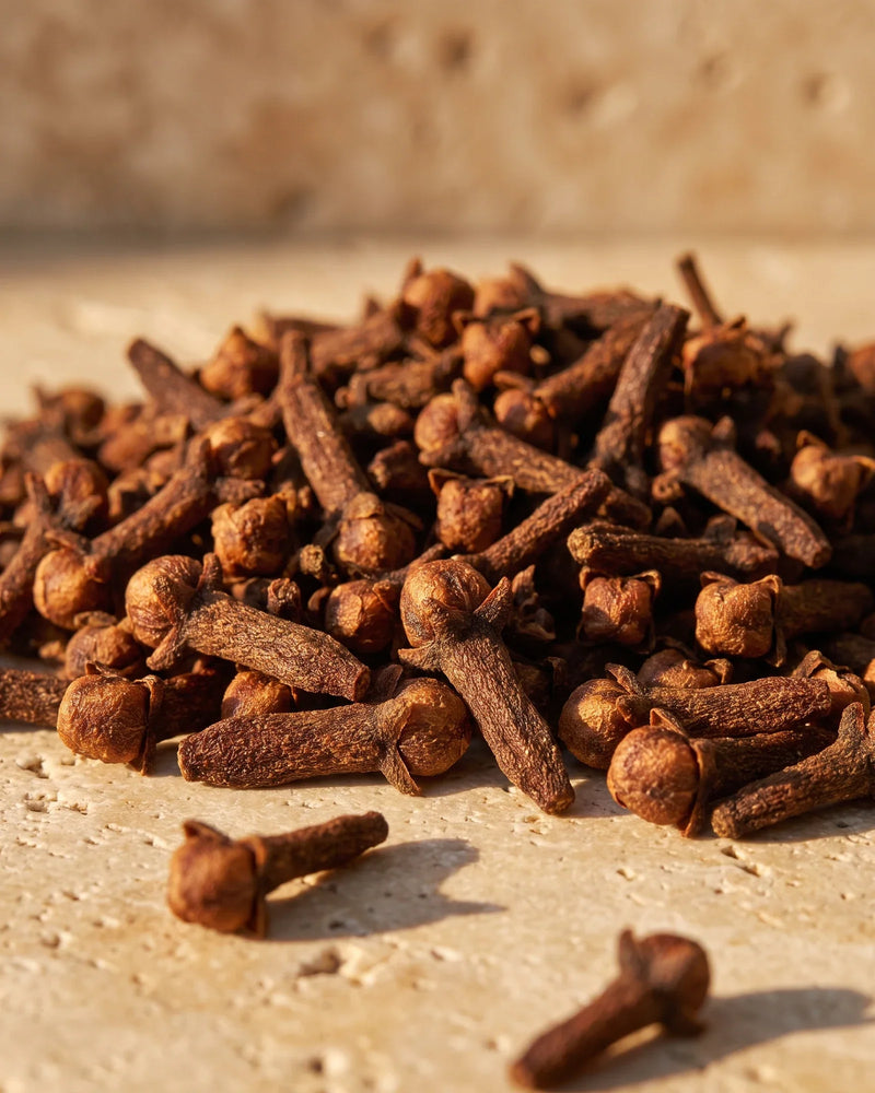 A heap of whole dried clove buds in raking warm light on travertine — the warm, spicy scent of Delune Clove Pure Essential Oil