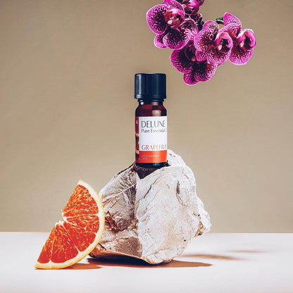 Grapefruit Pure Essential Oil bottle on rock with sliced grapefruit and purple flowers, promoting energy and healthy-looking skin care benefits.